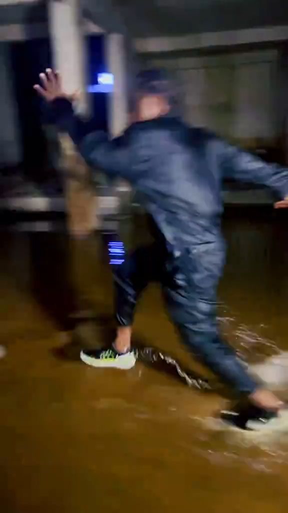 A vertical handheld smartphone video in a dark, flooded concrete basement or underground parking structure. Two men face off in ankle-dee...