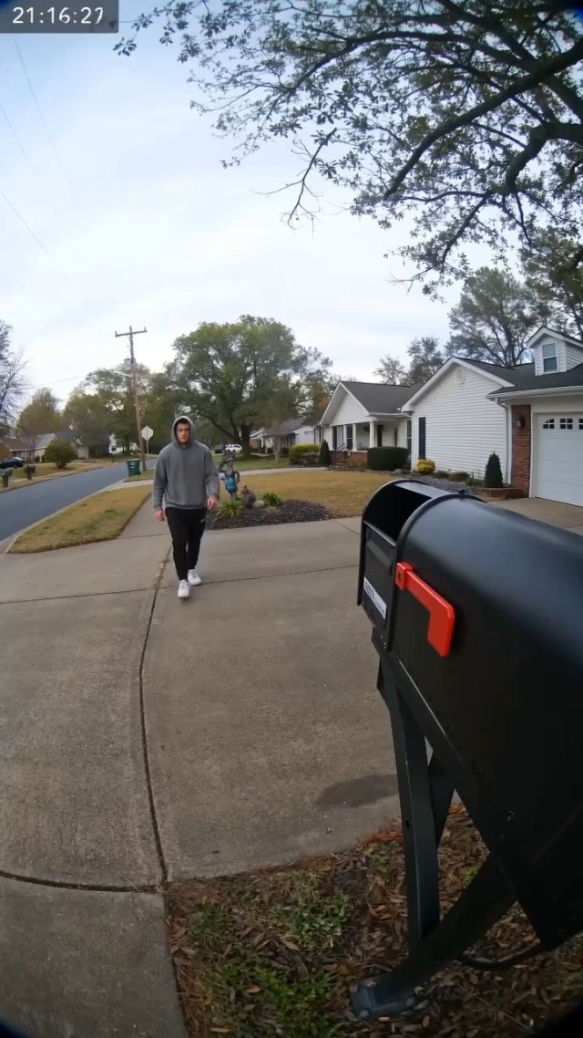 Security camera footage from a suburban driveway on a cloudy day, with a small white timestamp overlay in the top-left corner. A man in a...