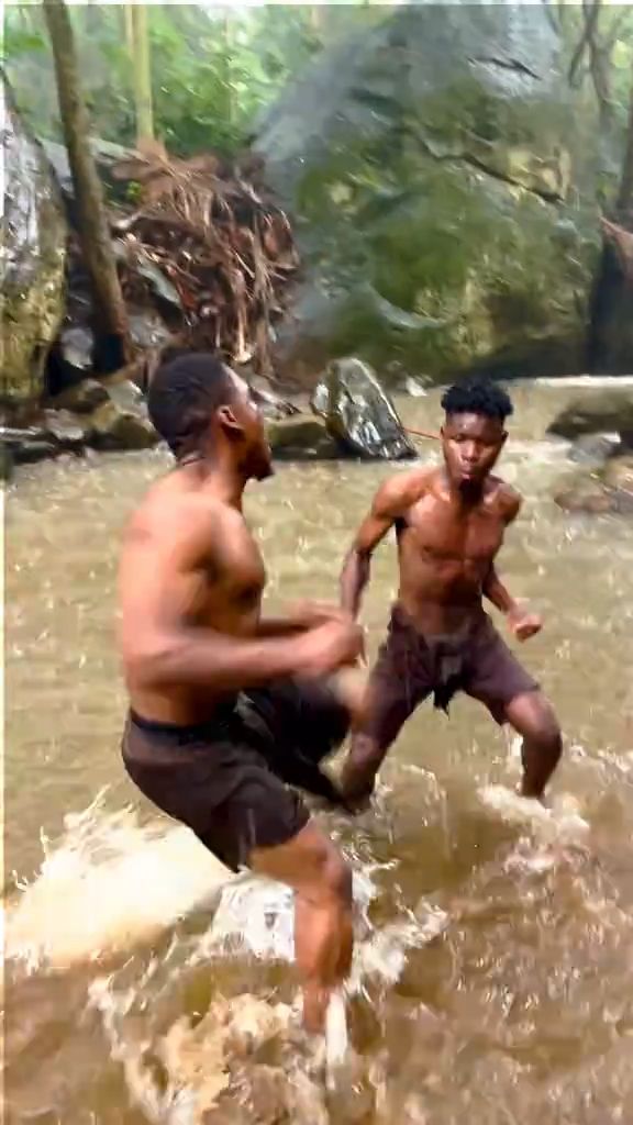 A vertical handheld smartphone video in a dense jungle creek during daylight. Two shirtless men fight in shallow muddy water surrounded b...