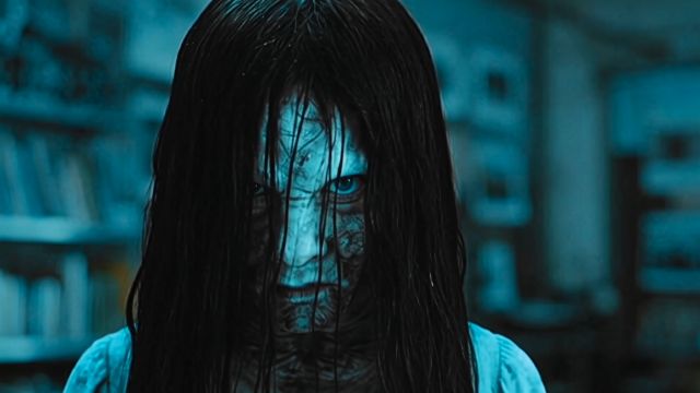 The woman, her long black hair obscuring her face, stands in a dimly lit room, her intense blue eyes locked onto the vi…