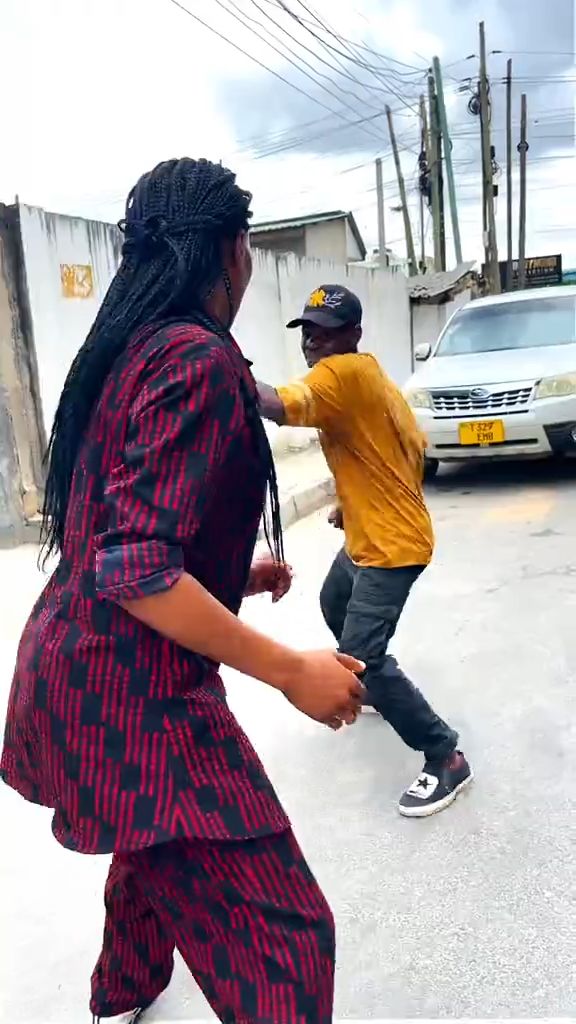 A vertical handheld smartphone video on a quiet urban side street during daylight. A woman in a red-and-black patterned outfit faces off ...