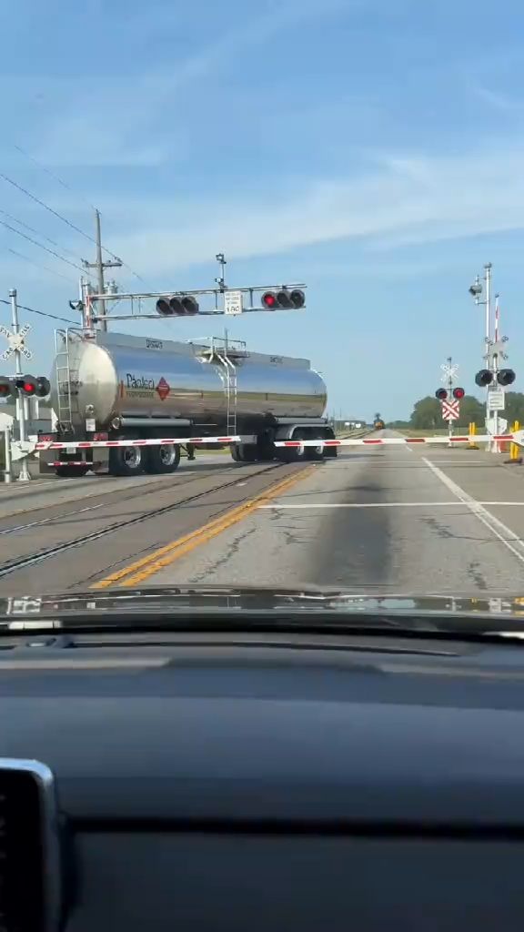 A vertical dashcam-style video shot from inside a stopped car at a rural railroad crossing in bright daylight. A silver fuel tanker truck...